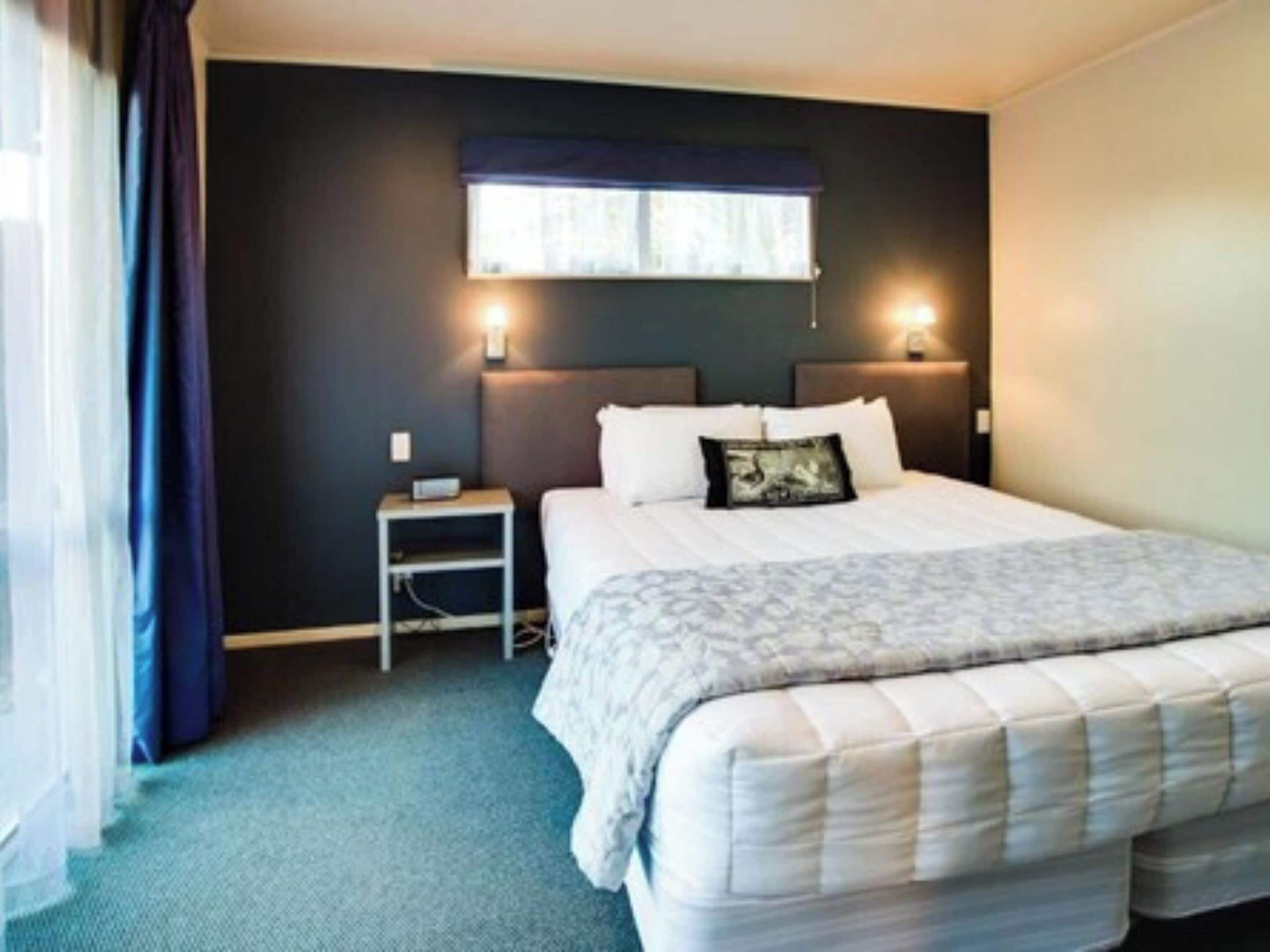 Une chambre d’hôtel avec un grand lit, une table de chevet, une lampe fixée au mur et une fenêtre avec des rideaux.