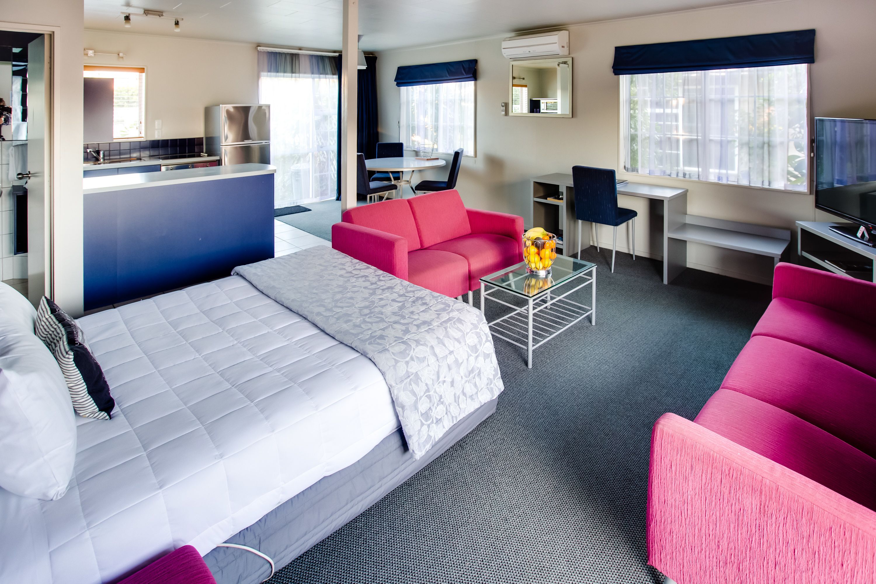 Een hotelkamer met een bed, een rode bank, een bureau met stoel, een televisie en een keukenhoek.