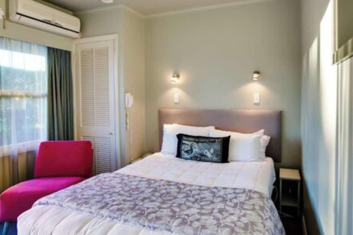 Una habitación de hotel con una cama, una silla roja, un equipo de aire acondicionado y dos lámparas fijadas a la pared.