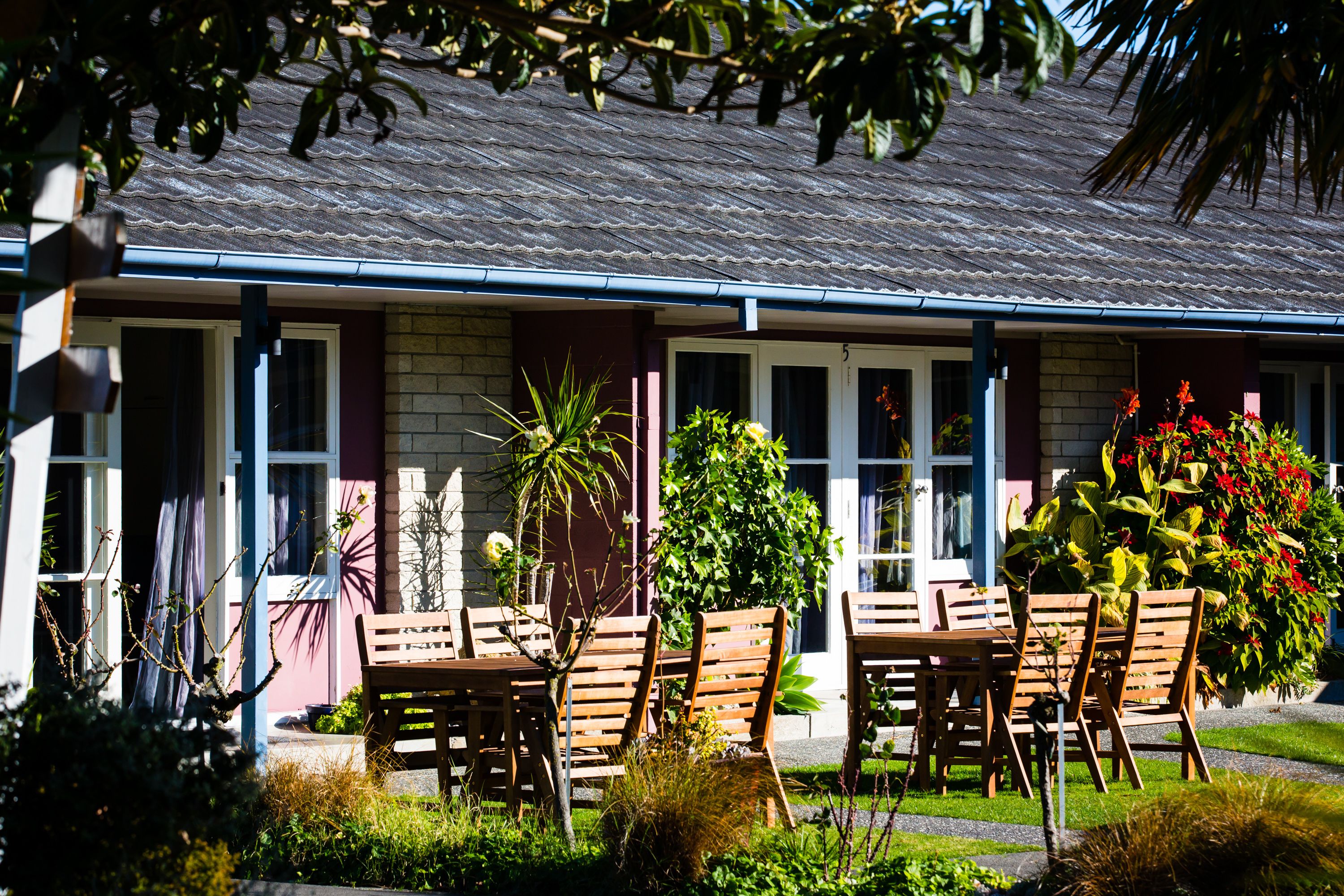 Un bâtiment avec un toit recouvert de tuiles, un porche à l’avant avec des chaises et une table en bois, et un jardin où poussent différentes plantes et fleurs.