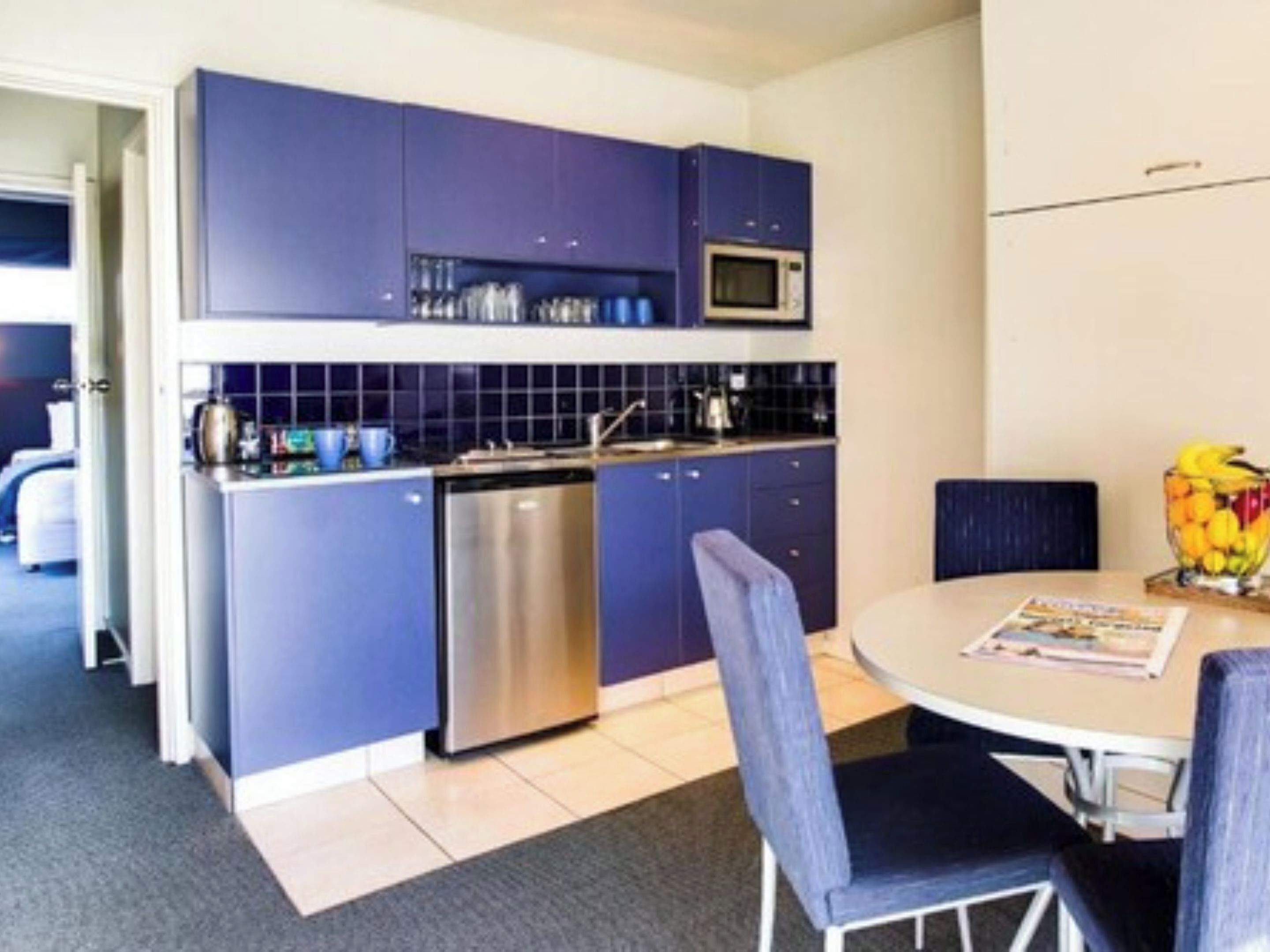 A modern kitchen with blue cabinets, a stainless steel dishwasher, and a microwave. There is a dining area with a round table and two chairs.