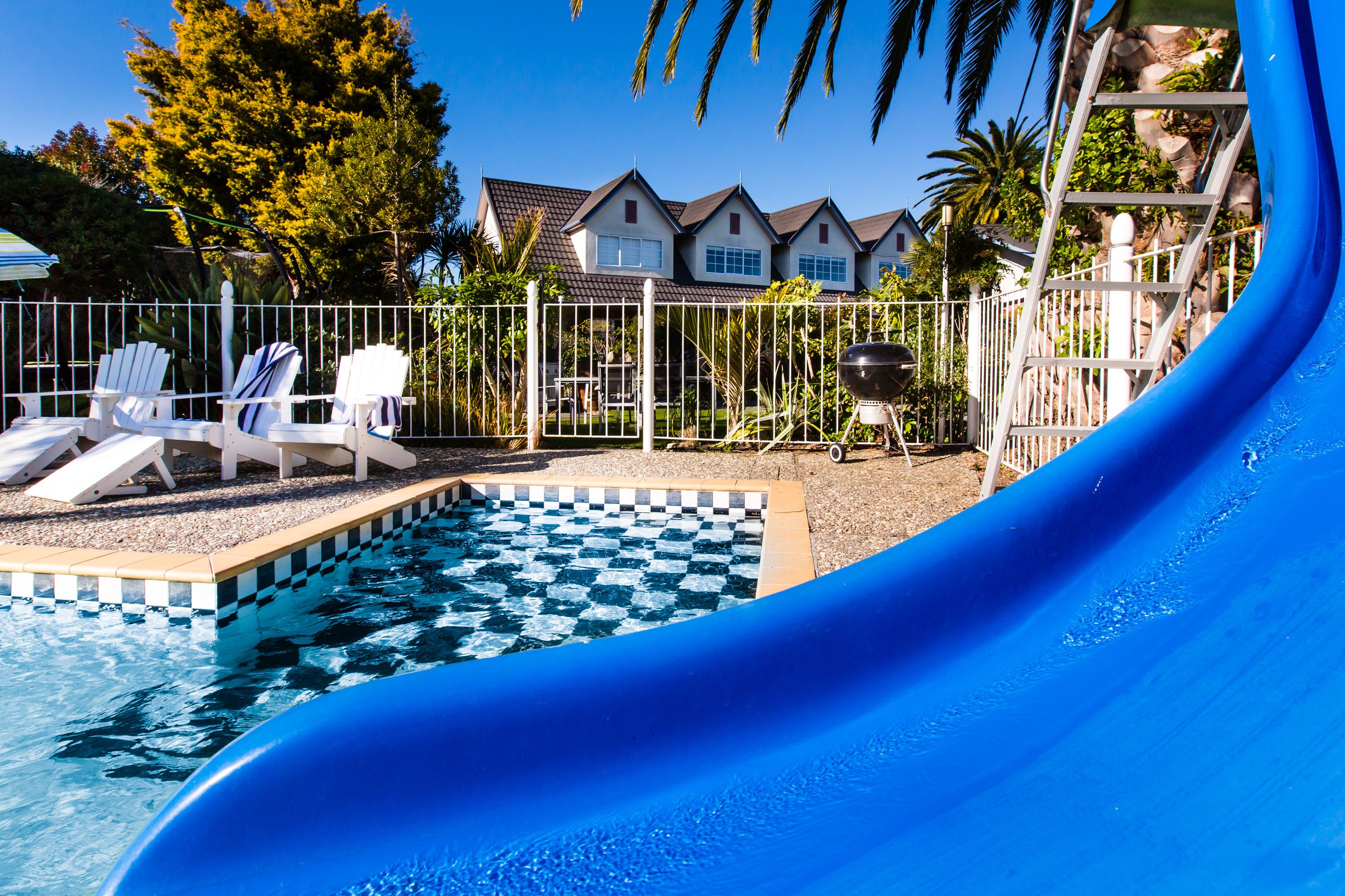 Piscina com escorregador azul, cadeiras de descanso brancas e área cercada com churrasqueira.
