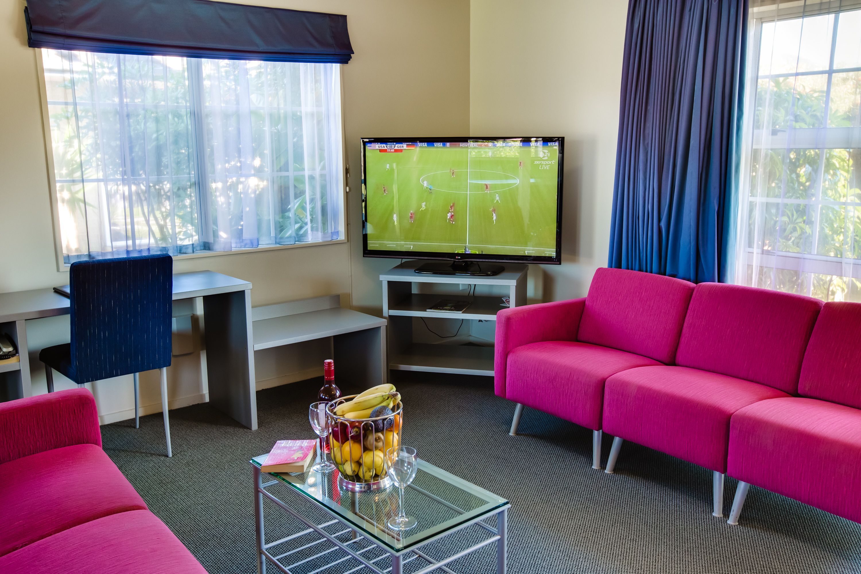 A living room with a TV displaying a sports game, a glass coffee table with a fruit bowl, a pink sofa, and a desk with a chair.