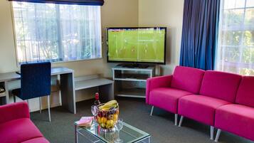 A living room with a TV displaying a sports game, a glass coffee table with a fruit bowl, a pink sofa, and a desk with a chair.
