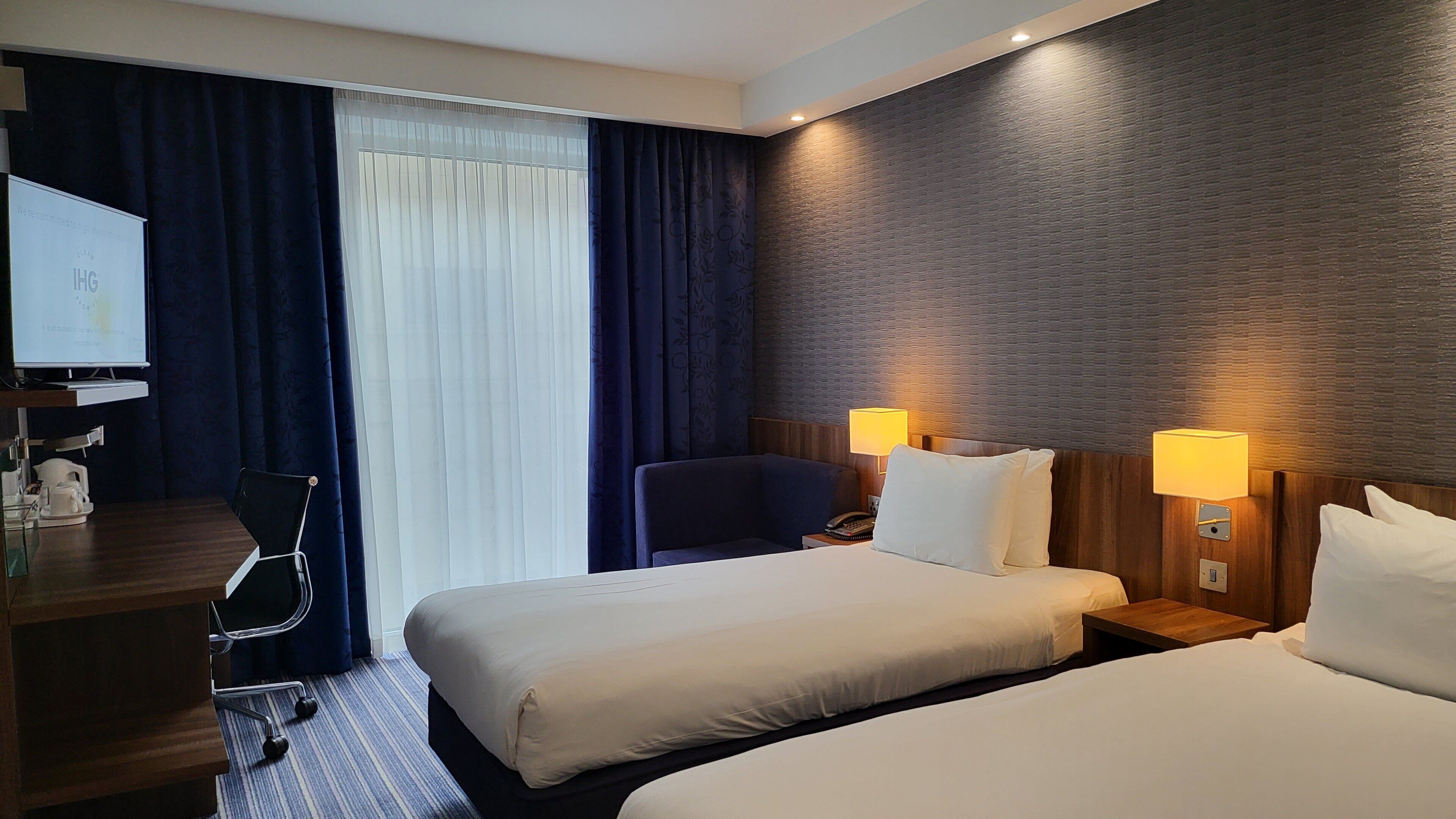 Ein Hotelzimmer mit zwei Betten, einem Schreibtisch mit Computer, einem Stuhl und einem fenster mit Vorhängen.