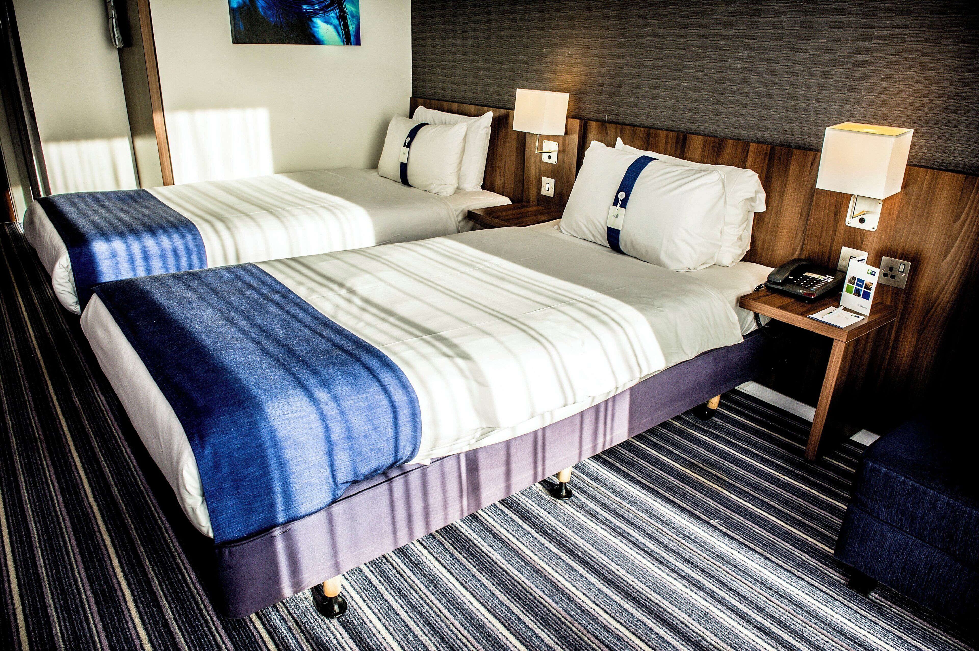Een hotelkamer met twee bedden, een houten hoofdbord, nachtlampjes, een telefoon en een klein tafeltje met een brochure.