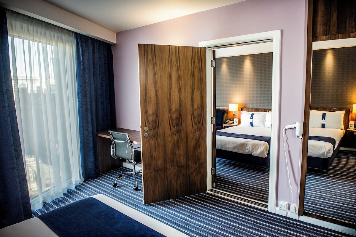 Quarto de hotel com cama, escrivaninha, cadeira, espelho e janela com cortinas.