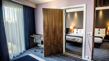 Une chambre d’hôtel avec un lit, un bureau, une chaise, un miroir et une fenêtre avec des rideaux.