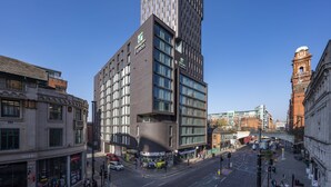 A modern hotel with a prominent 'Express' sign, a pedestrian crossing, and a historic clock tower in the background. - Holiday Inn Express Manchester CC - Oxford Road by IHG (Manchester)