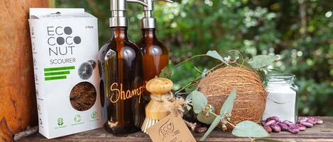 A wooden shelf with a box of ECO COCO NUT SCOURER, two bottles of shampoo, a coconut, a jar of white powder, and a wooden brush.