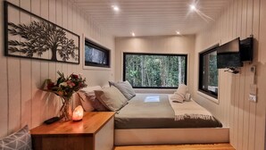 Een slaapkamer met een bed, een nachtkastje, een vaas met bloemen, een zoutlamp en een groot raam met uitzicht op een bos.