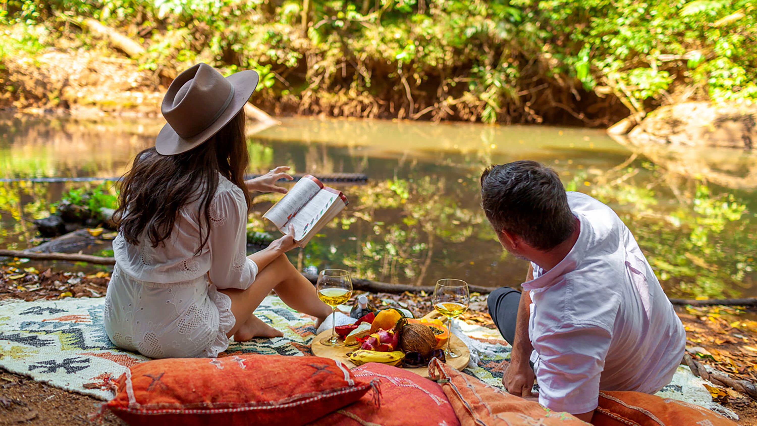Dos personas sentadas sobre una manta junto a un cuerpo de agua tranquilo, leyendo un libro.