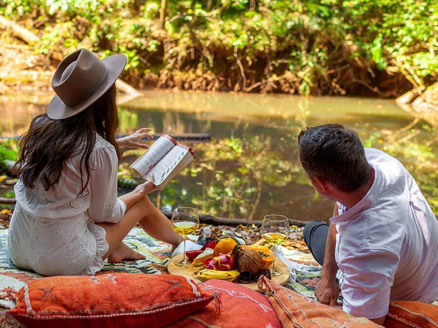Two people sitting on a blanket by a calm body of water, reading a book.