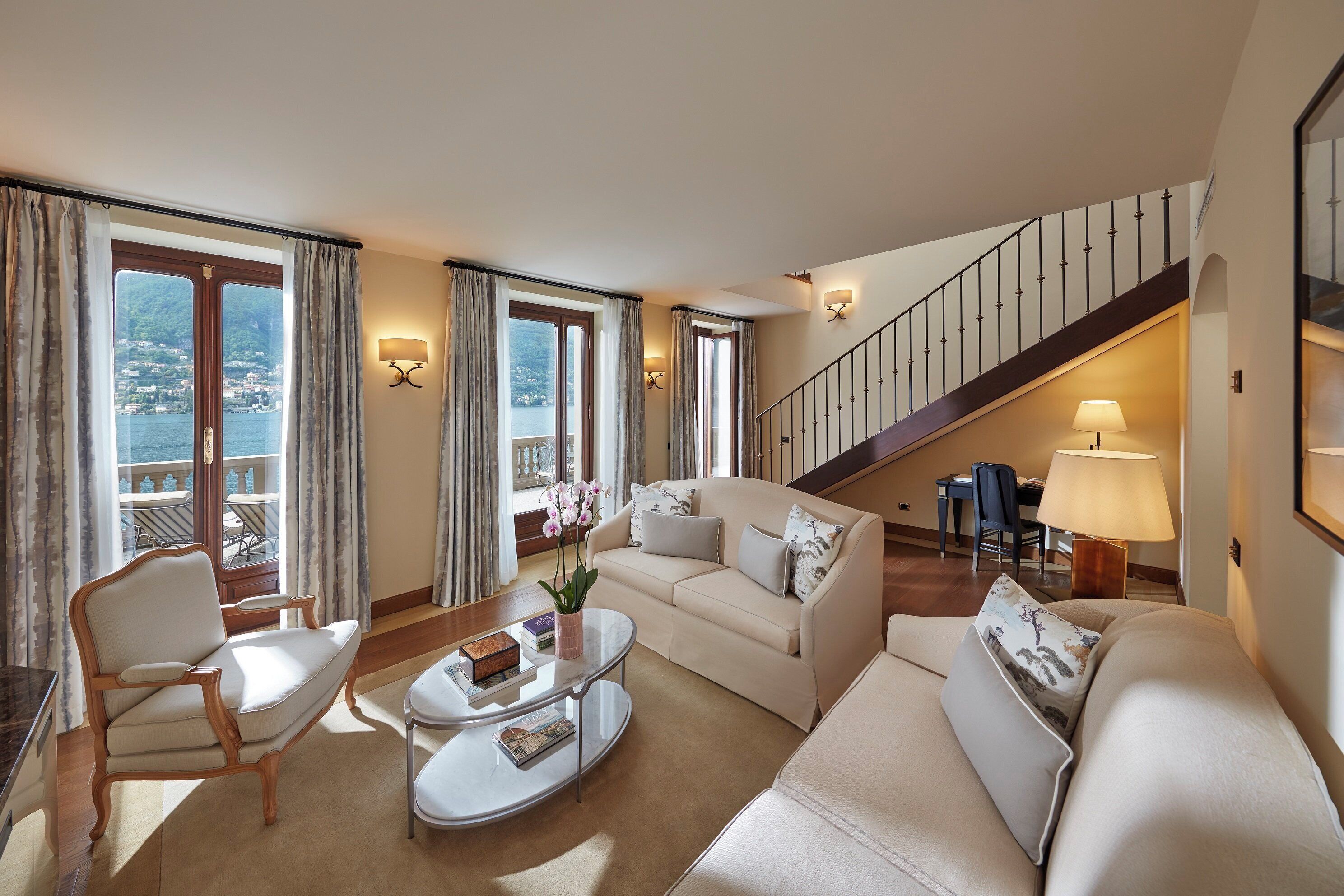 A spacious living room with a sofa, armchair, and coffee table. A staircase leads to a balcony with a view of the water.