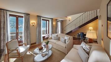 A spacious living room with a sofa, armchair, and coffee table. A staircase leads to a balcony with a view of the water.
