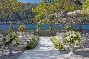 An outdoor wedding aisle with white flowers and chairs, overlooking a lake and town.