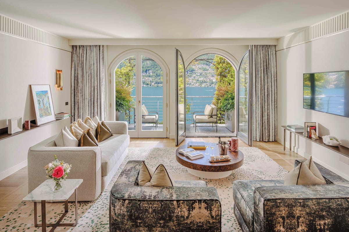 Un salón amplio con un sofá grande, una mesa de centro y un televisor montado en la pared. Dos ventanas amplias con cortinas ofrecen vistas a un lago y vegetación.