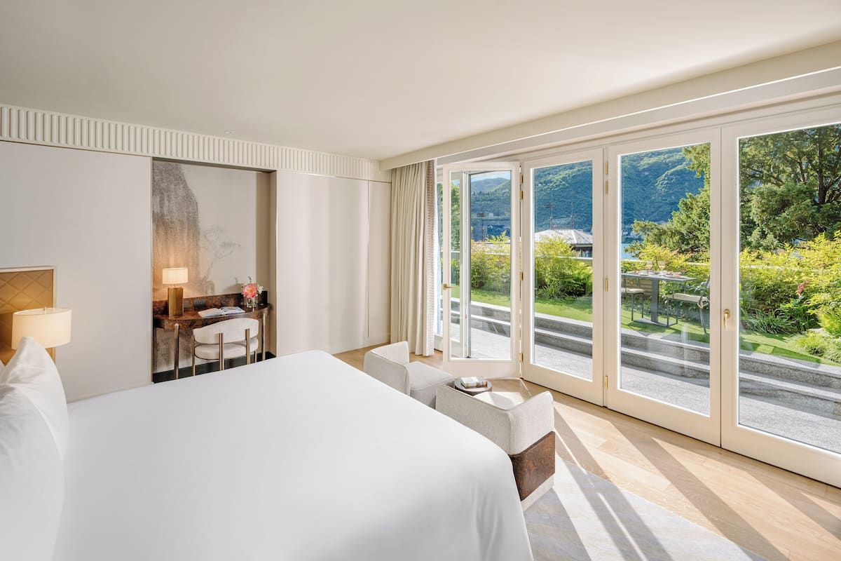 Habitación de hotel con una cama grande, un área de descanso con una mesa, un escritorio con una lámpara y vistas a un paisaje montañoso a través de puertas correderas amplias.