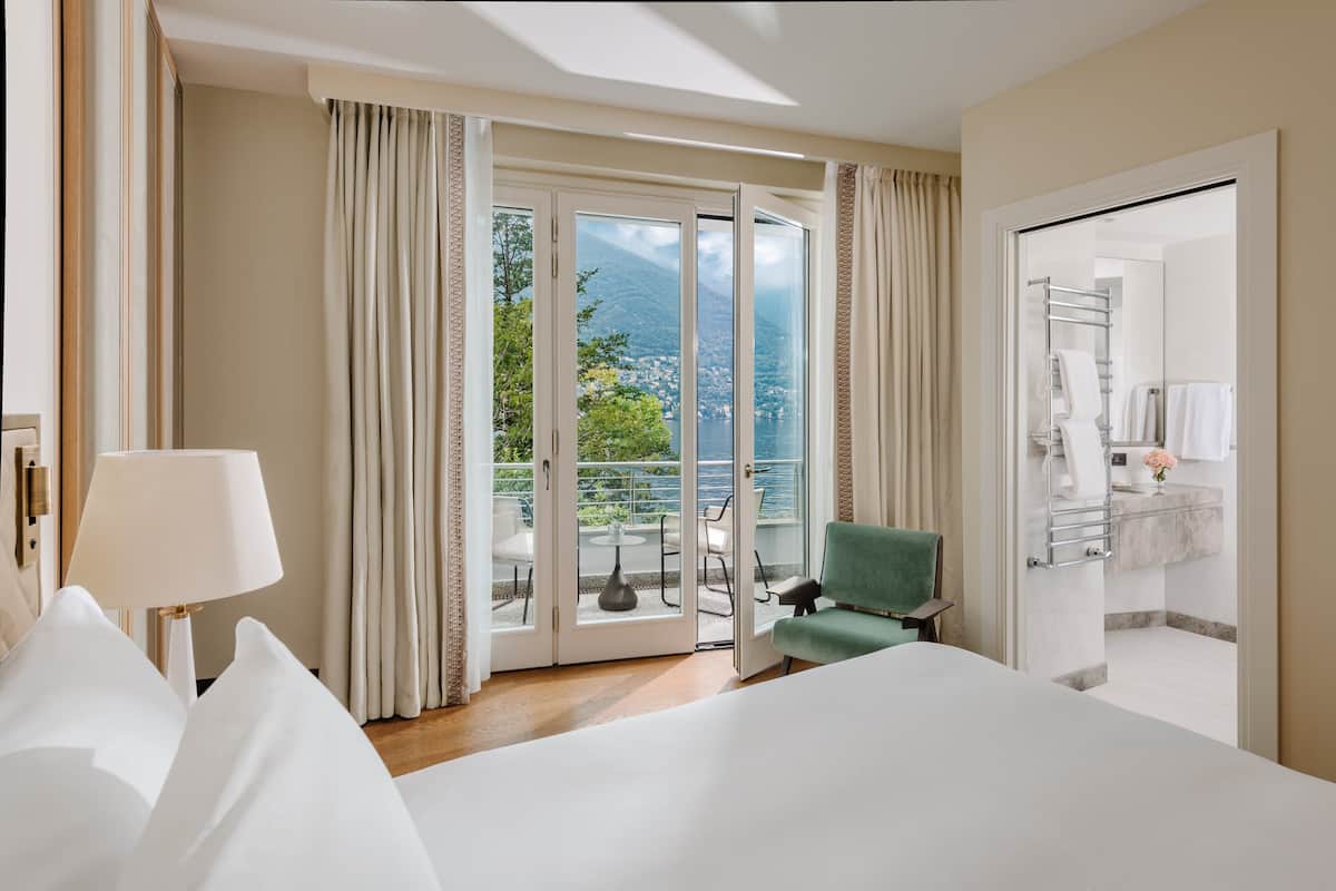 Habitación de hotel con una cama grande, un sillón verde, un balcón con vistas a las montañas y un baño con toallero.