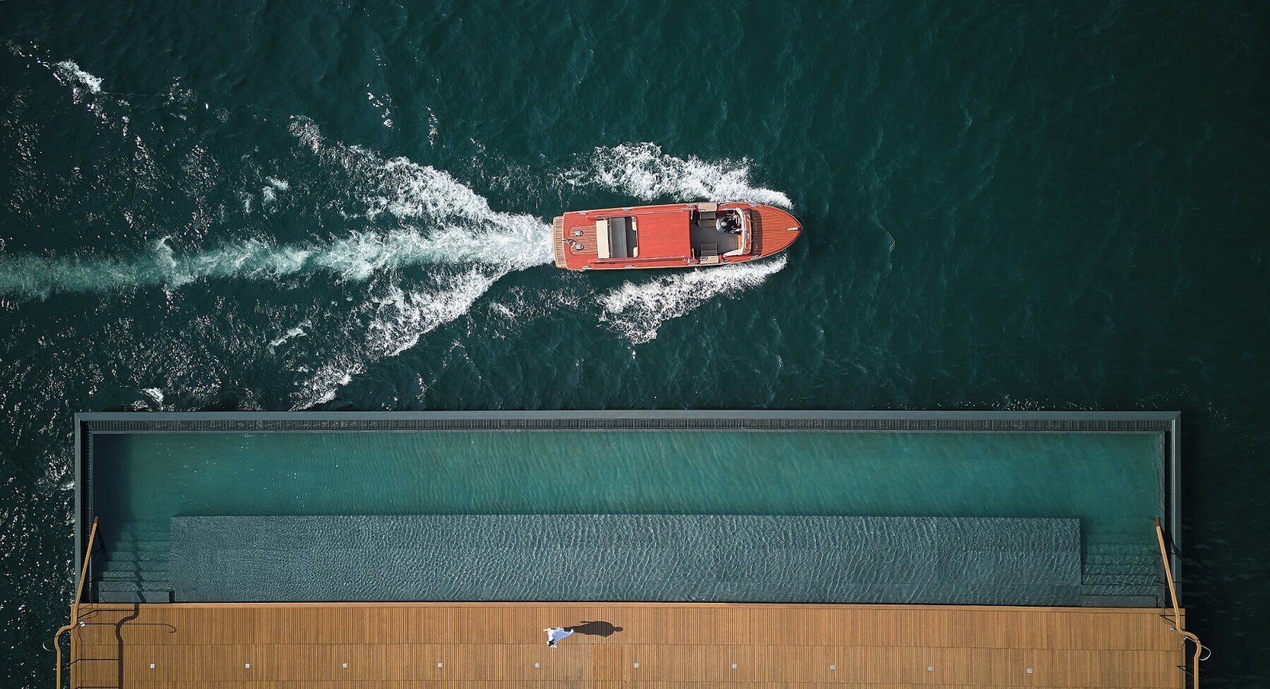 A boat on a calm body of water, a dock with a wooden deck, and a person standing on the dock.