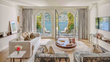 A spacious living room with a large sofa, a coffee table, and a TV mounted on the wall. There are two large windows with curtains, offering a view of a lake and greenery.