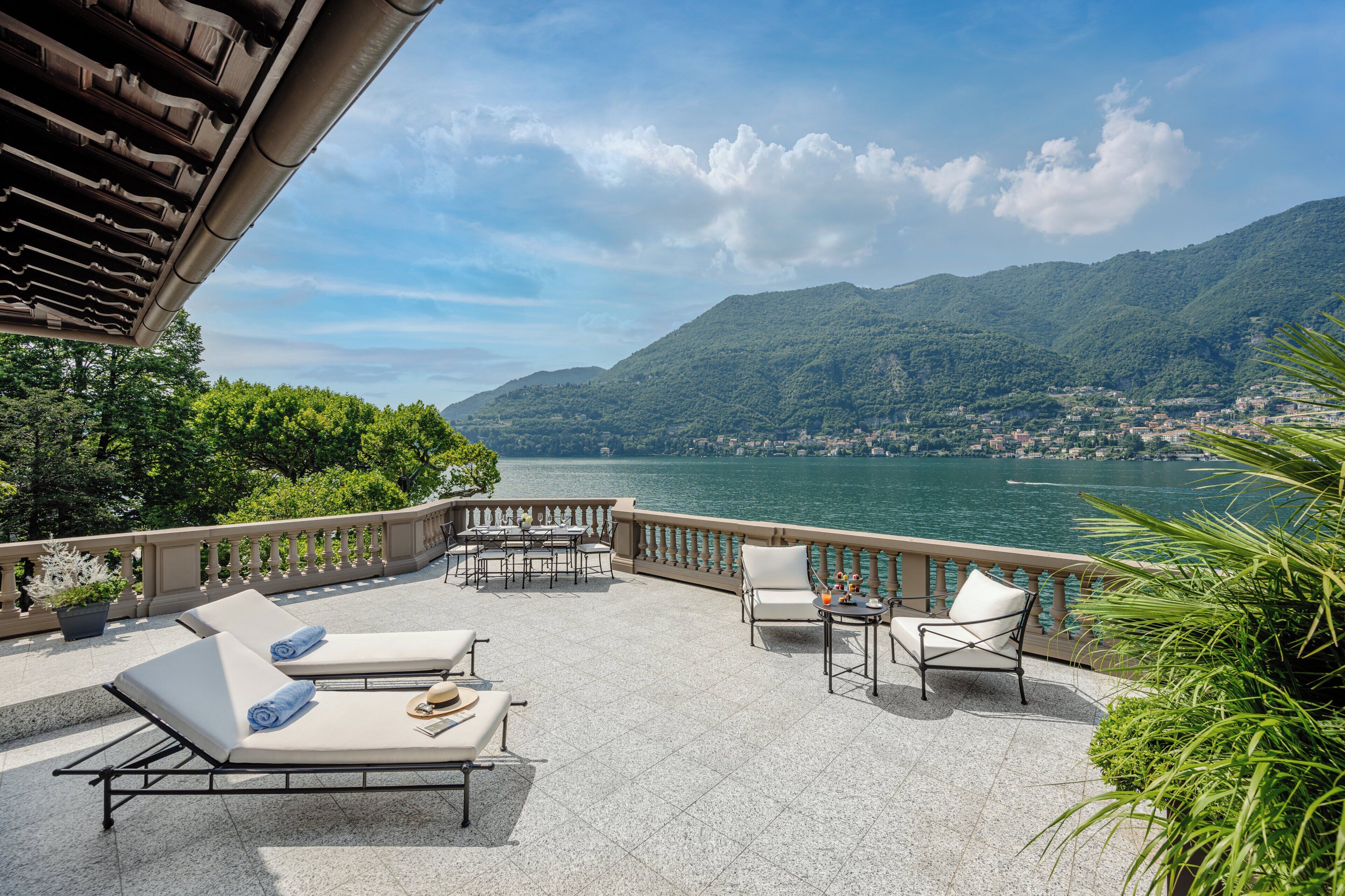 A terrace with lounge chairs and a table, overlooking a lake and mountains.