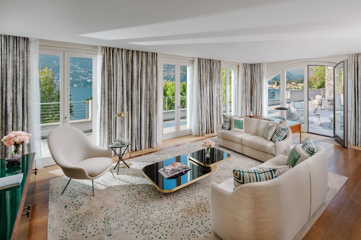 Una sala de estar moderna con sofá, sillón y mesa de centro, ventanas amplias con cortinas, y vistas al agua y a las montañas.