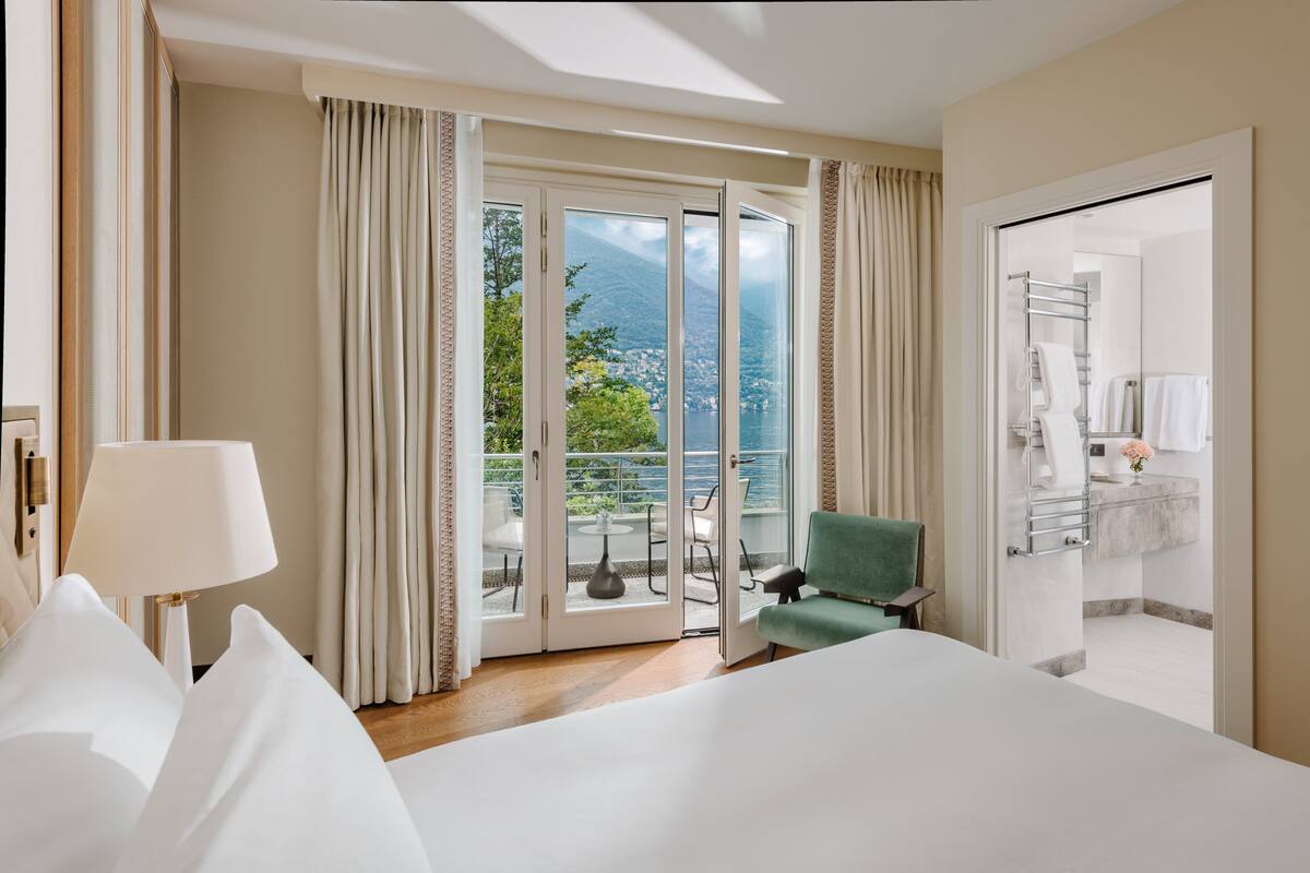 A hotel room with a large bed, a green armchair, a balcony with a view of the mountains, and a bathroom with a towel rack.