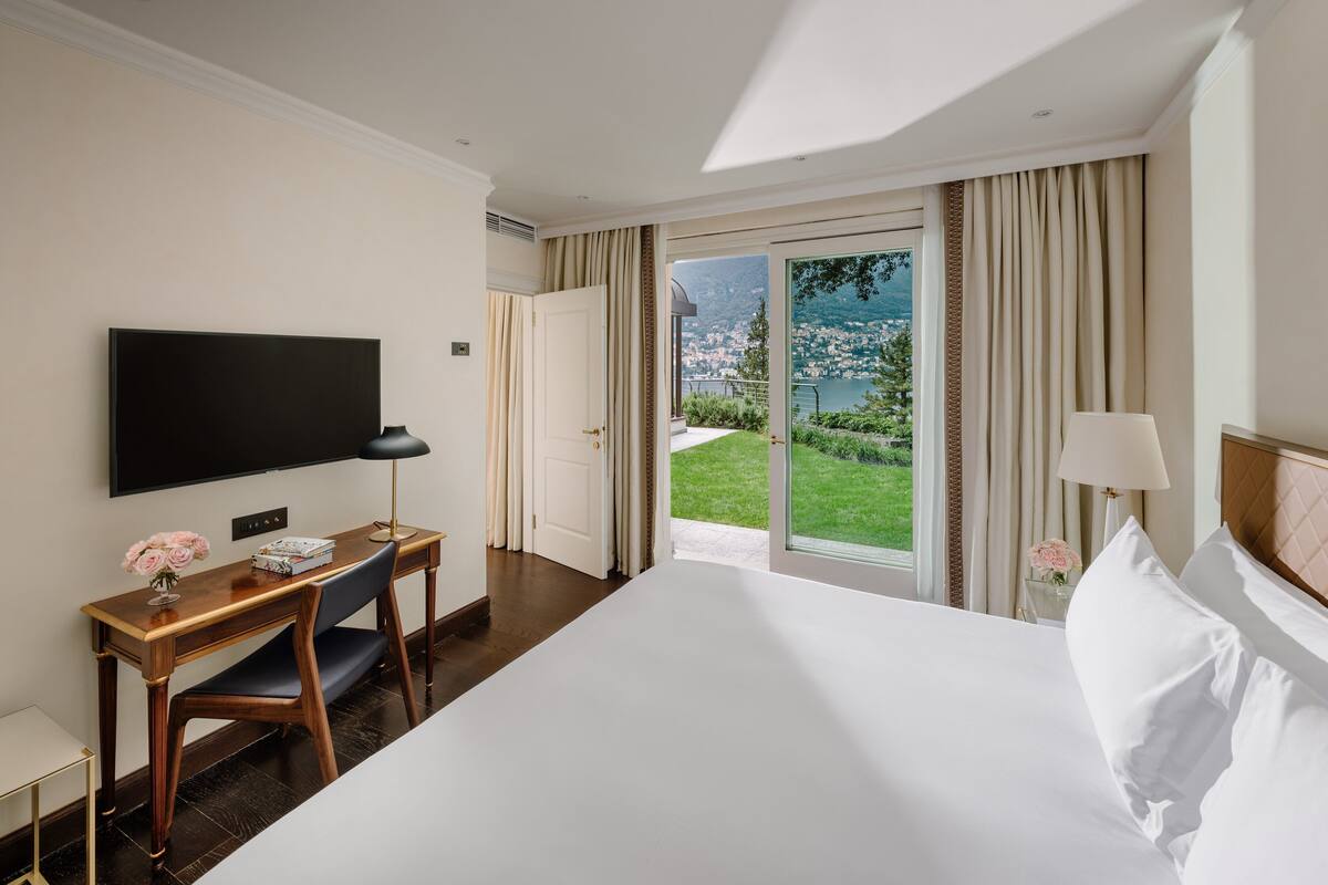 Habitación de hotel con una cama grande, un escritorio con una silla, un televisor de pantalla plana y vistas a una ciudad y zonas verdes a través de una puerta corrediza de cristal.