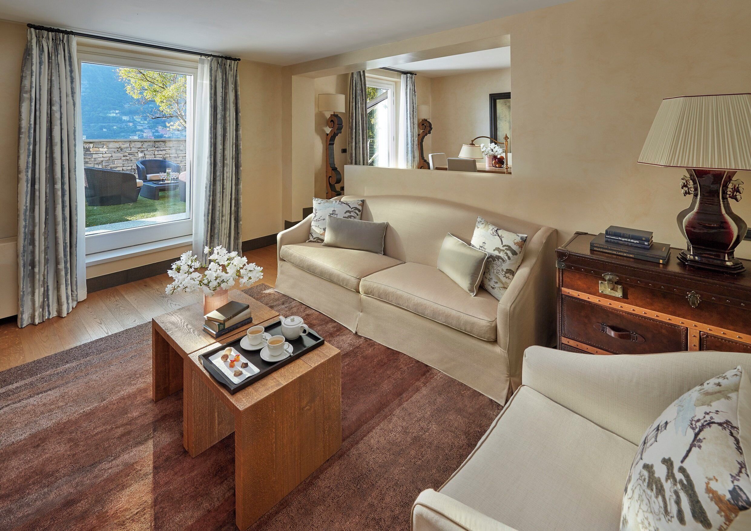 Ein Hotelzimmer mit einer Couch, einem Couchtisch mit einer Auswahl an Gebäck, einem Holznachttisch, einer Lampe und Blick auf die Berge durch das Fenster.