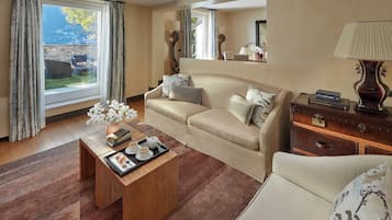 Ein Hotelzimmer mit einer Couch, einem Couchtisch mit einer Auswahl an Gebäck, einem Holznachttisch, einer Lampe und Blick auf die Berge durch das Fenster.