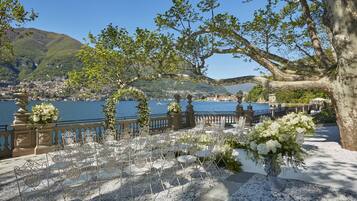 Une cérémonie de mariage en plein air, avec des chaises et des tables blanches, une arche décorative et une vue sur un lac et des montagnes.