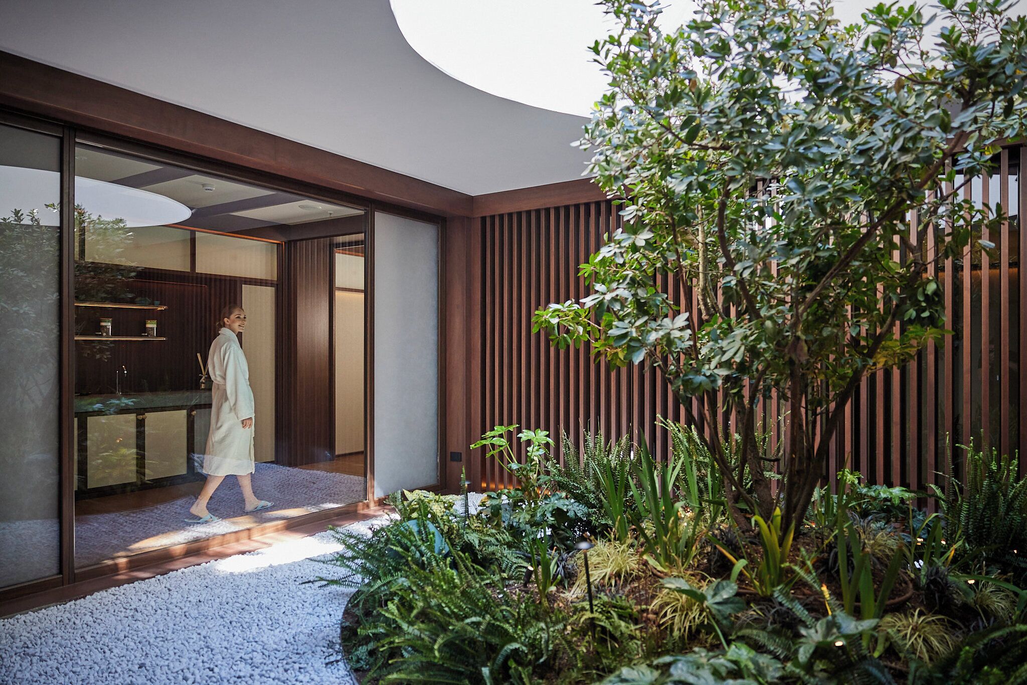 A modern building with a glass entrance, a wooden interior, and a garden with various plants.