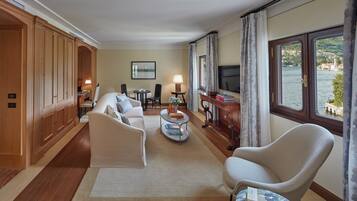 Habitación de hotel con sofá, sillones, una mesita, televisor y vista a un lago.