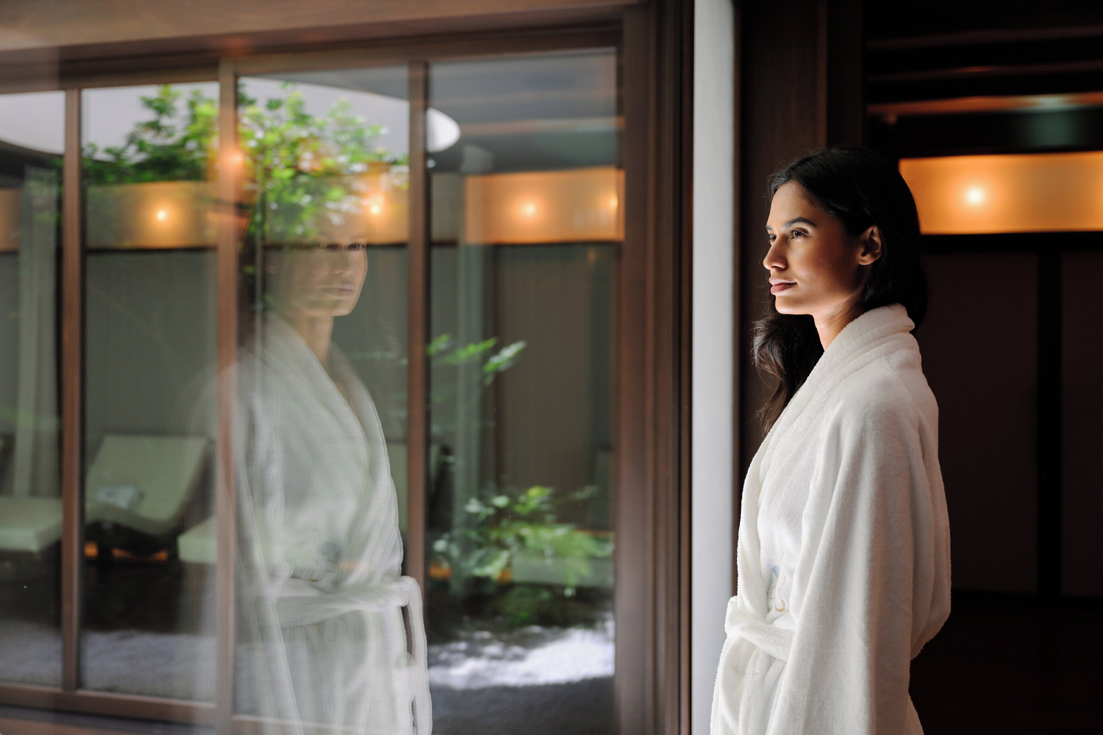 A person in a white bathrobe standing by a glass door with a view of a garden and outdoor seating.