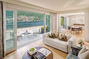 Una sala de estar moderna con sofá, mesa de centro y comedor. Un amplio ventanal da a un balcón con vistas panorámicas a un lago y colinas.