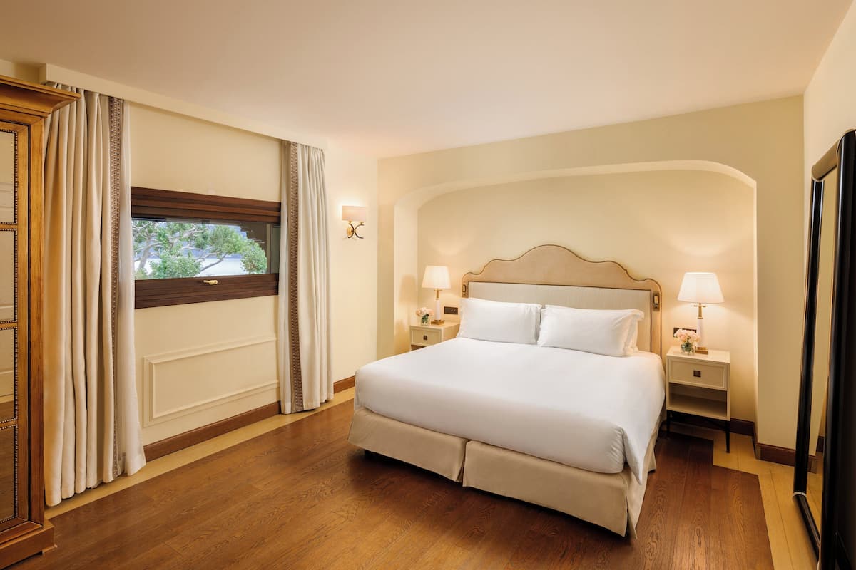 Habitación de hotel con una cama grande, dos mesitas de noche con lámparas, un ventanal con cortinas y suelo de madera.