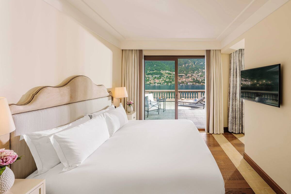 A hotel room with a large bed, a TV, and a balcony with a view of a town and water.
