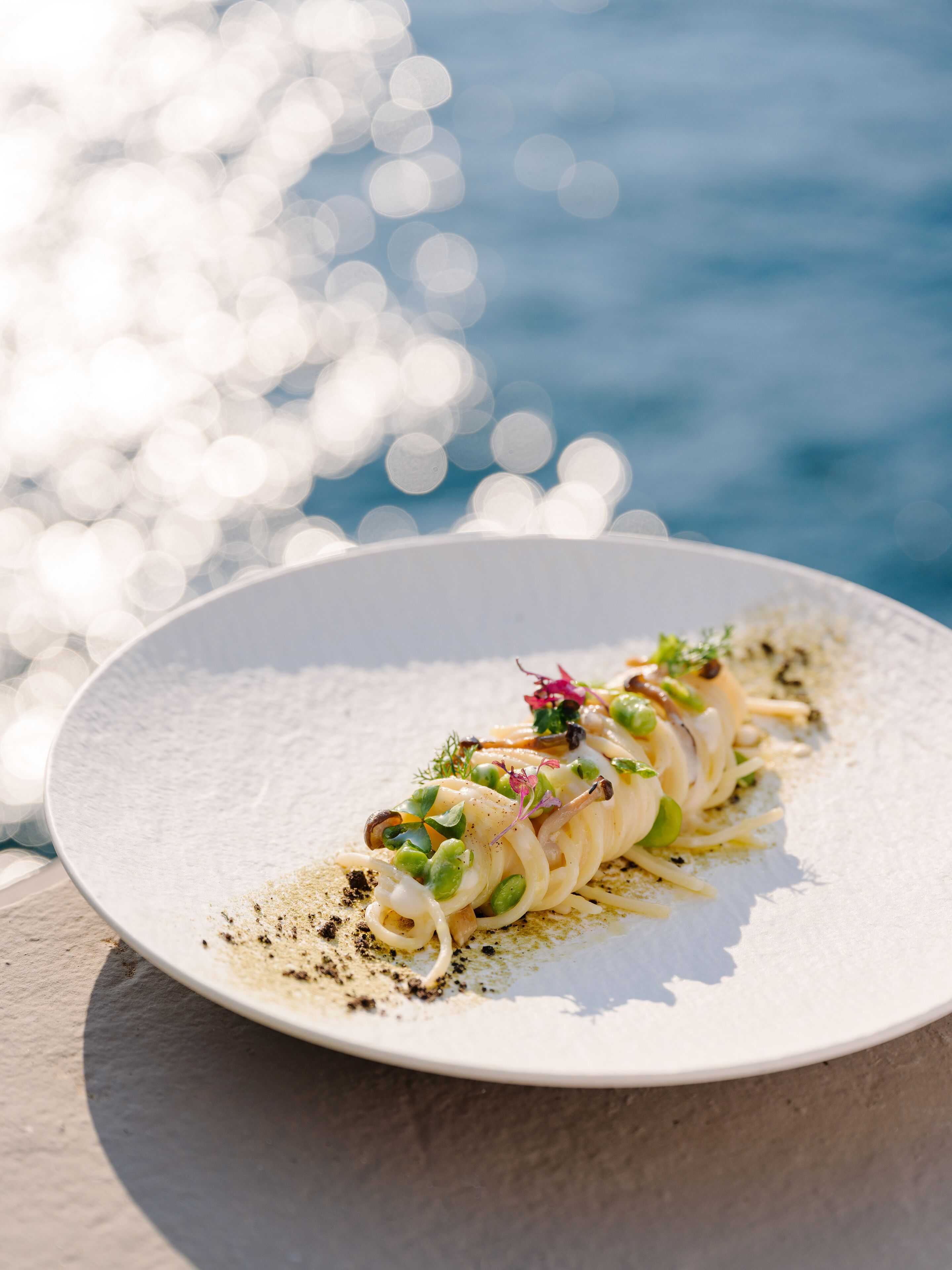 Une assiette blanche contenant un plat de fruits de mer, agrémenté d’herbes et de fleurs comestibles, posée sur une surface texturée près d’une étendue d’eau, avec des lumières floues en arrière-plan.