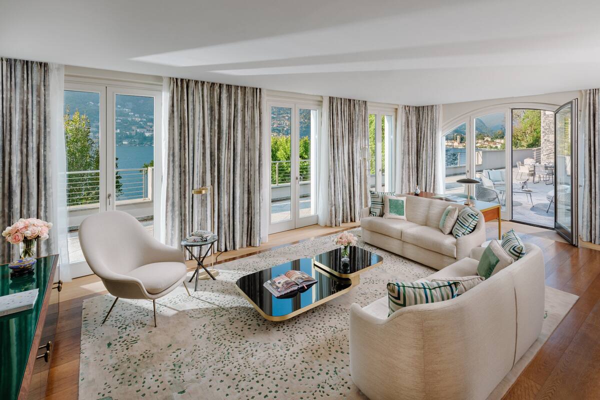 A modern living room with a sofa, armchair, and coffee table, large windows with curtains, and a view of a body of water and mountains.