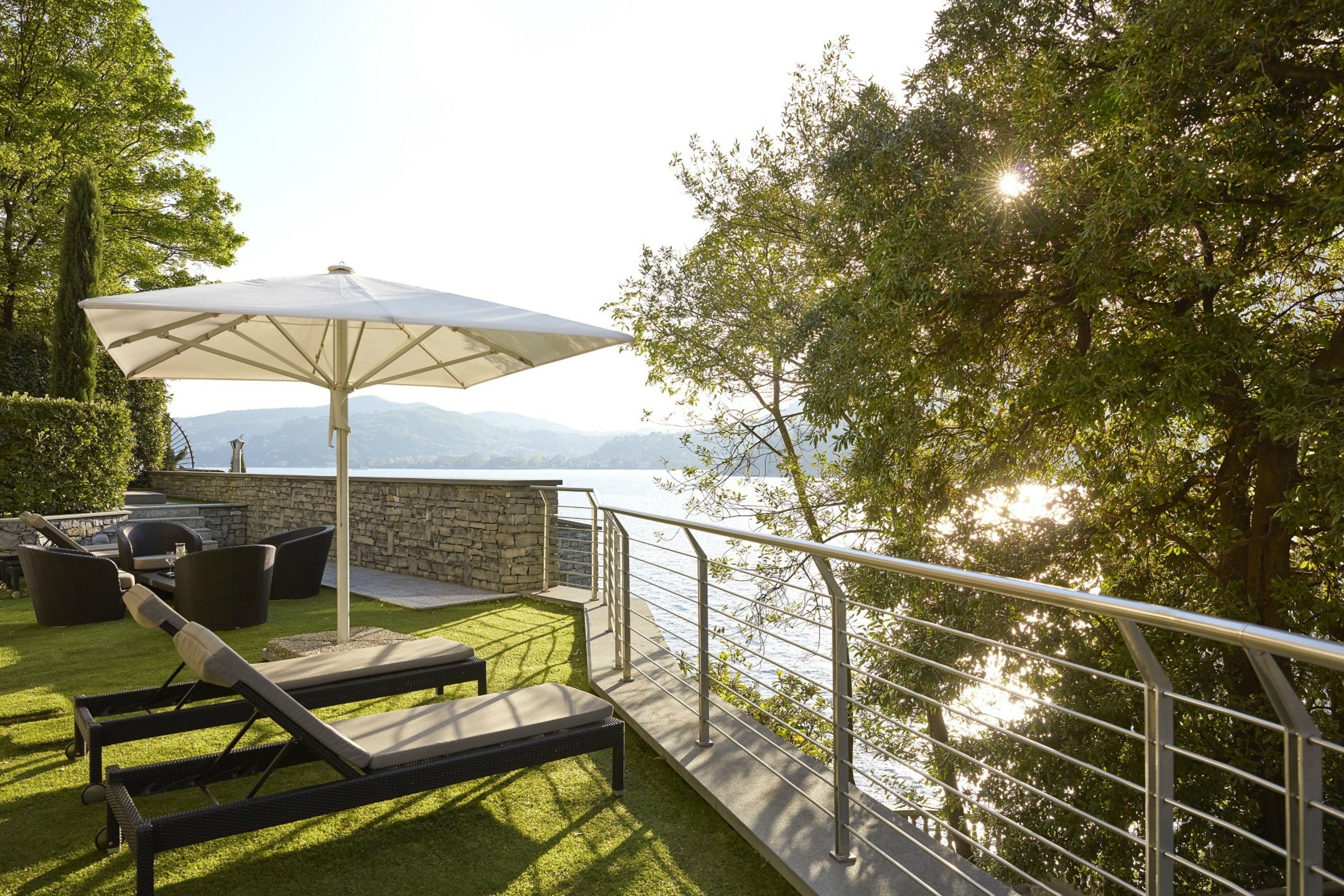 Eine Terrasse im Freien mit einer Steinmauer, einem weißen Sonnenschirm und Blick auf den See.