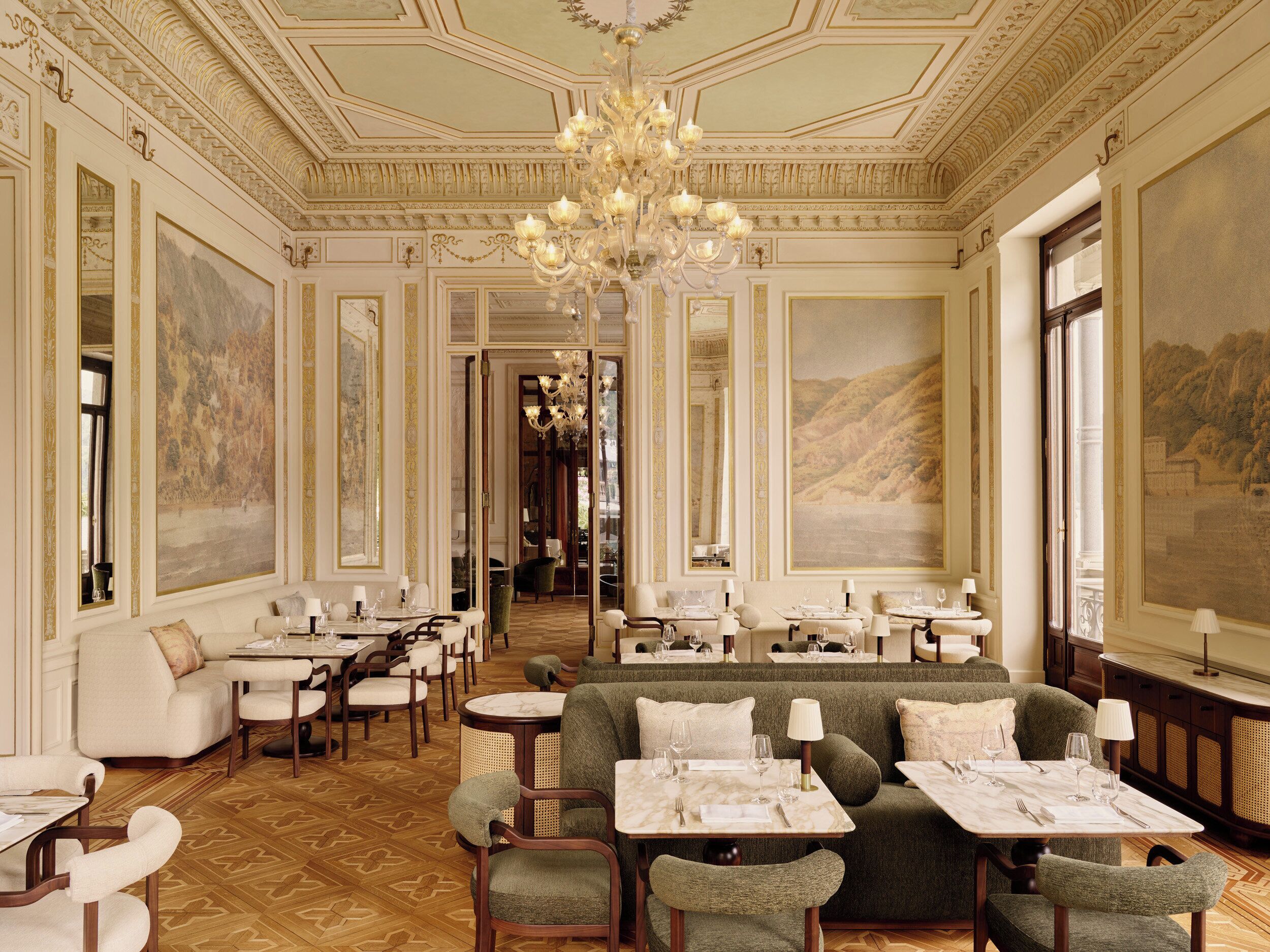 A luxurious hotel lobby with ornate ceiling details, a grand chandelier, and a cozy seating area with tables set for dining.