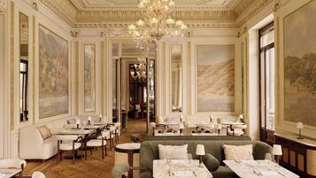 A luxurious hotel lobby with ornate ceiling details, a grand chandelier, and a cozy seating area with tables set for dining.