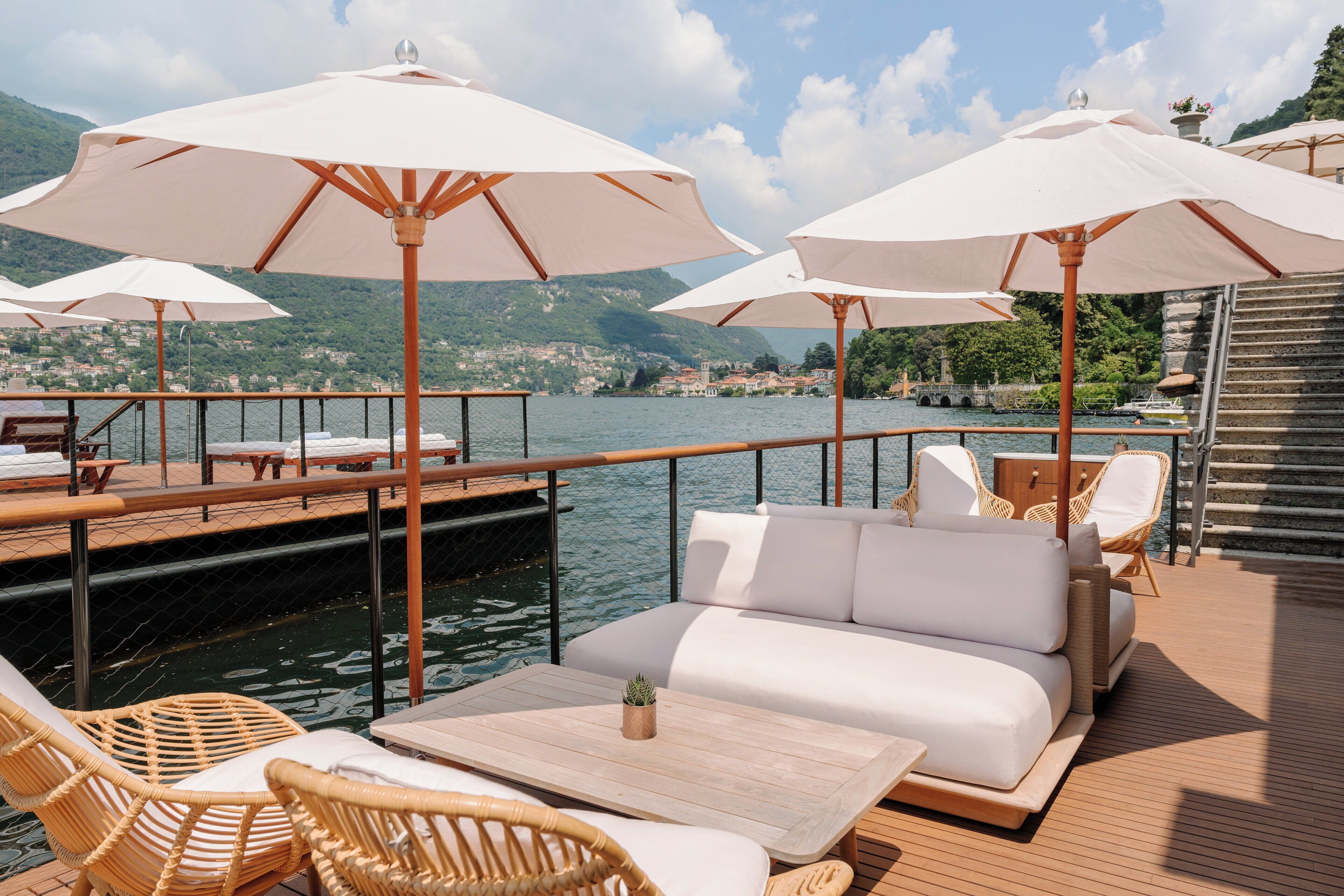 A deck with white outdoor furniture, including a sofa and chairs, under large umbrellas, overlooking a body of water with a mountainous backdrop.
