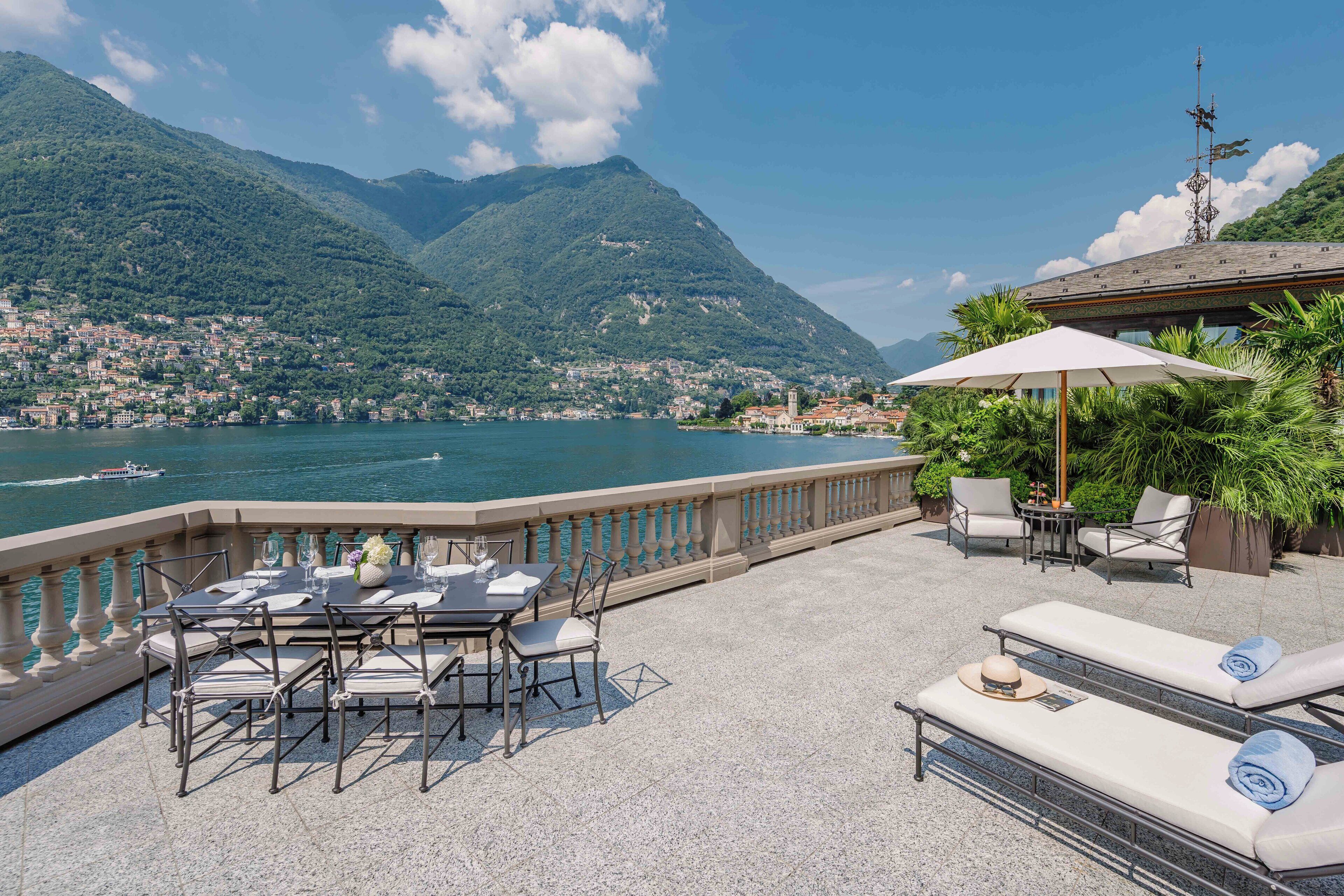 Una terrazza con posti a sedere all'aperto, un tavolo apparecchiato per la cena e vista su un lago con montagne sullo sfondo.