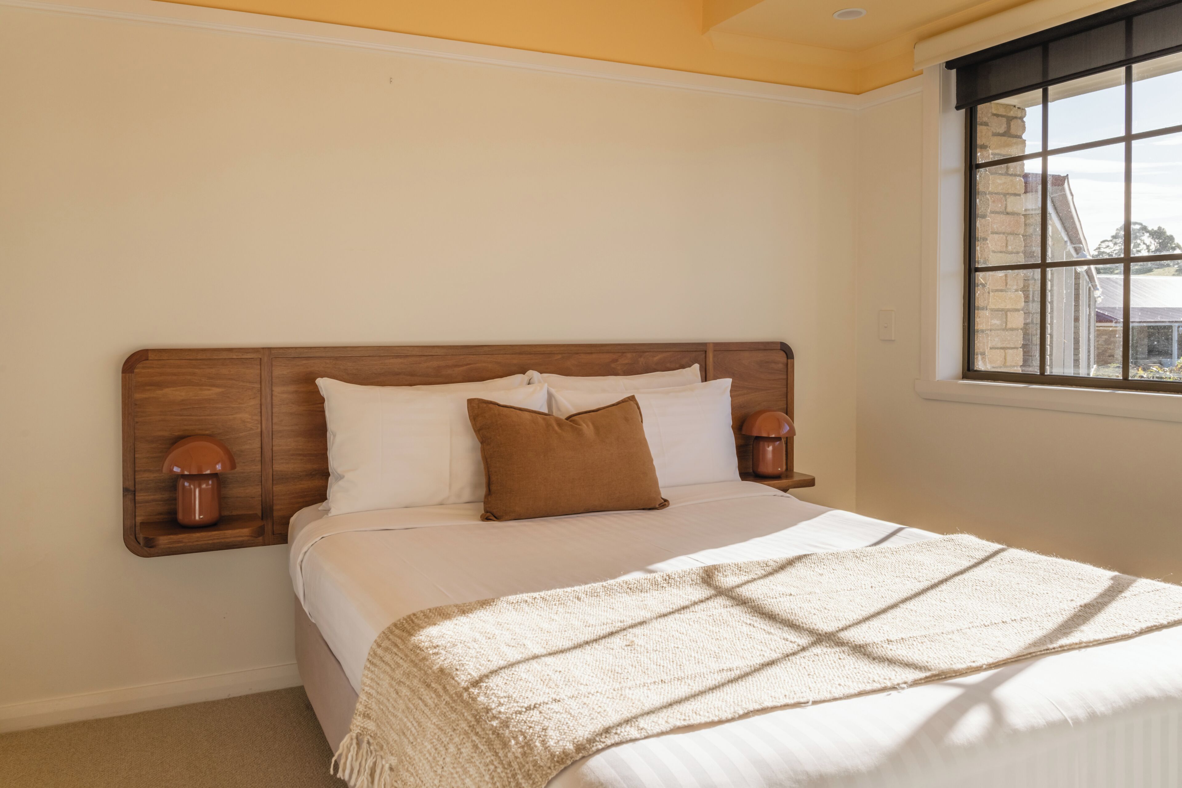 A bedroom with a wooden headboard, a bed with white bedding, two bedside lamps, and a window with a view of buildings.