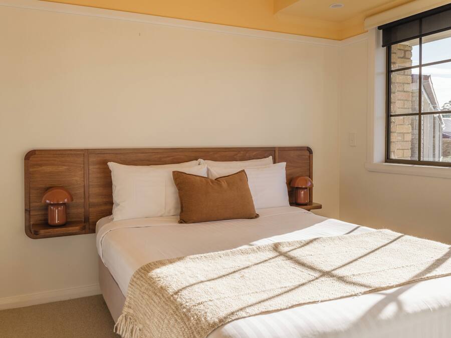A bedroom with a wooden headboard, a bed with white bedding, two bedside lamps, and a window with a view of buildings.