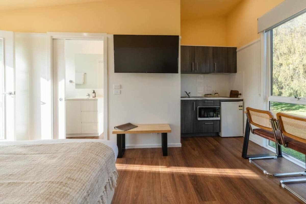 A bedroom with a bed, a small table, a bench, a TV, a kitchenette, and a window with a view.