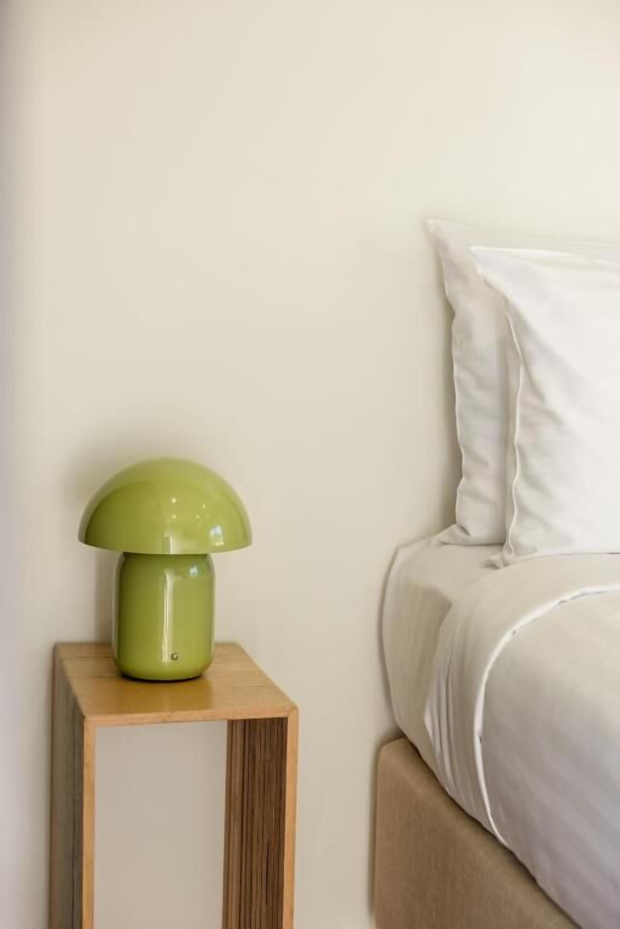 A bed with white bedding, a green lamp on a wooden nightstand, and a plain wall.