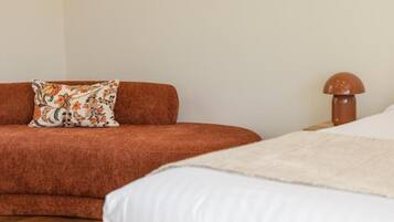 A room with a brown sofa, a patterned pillow, a white bed, and a wooden nightstand with a lamp.